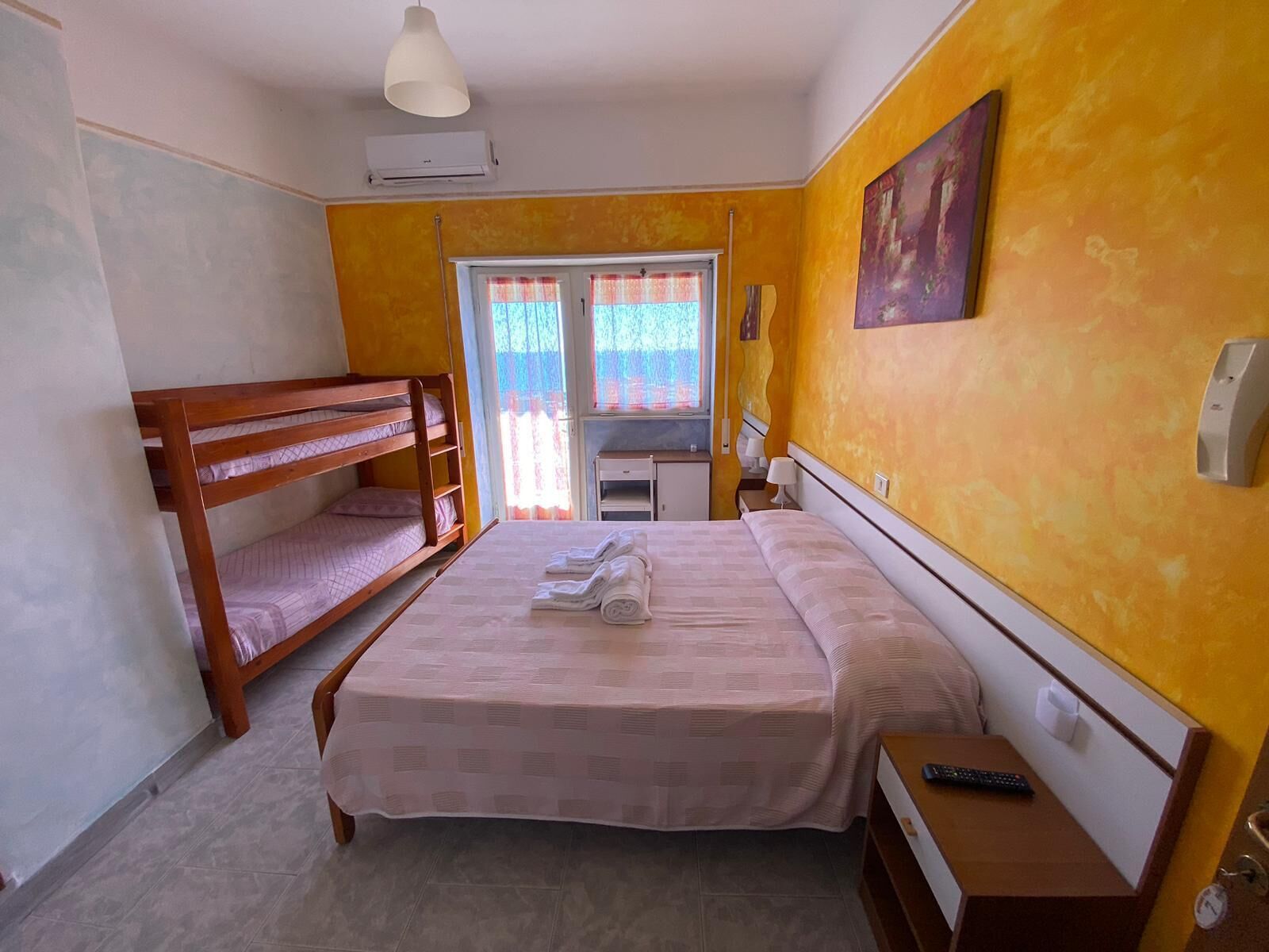 Una stanza con un letto a castello, un letto singolo, una scrivania, una sedia, una lampada e un quadro appeso al muro.