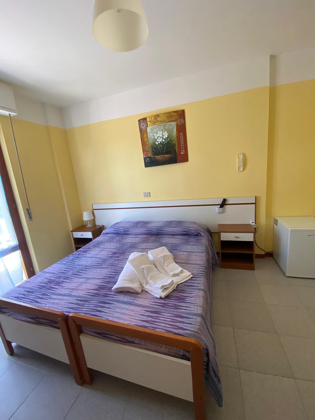 A bedroom with a bed, a nightstand, a lamp, a wall-mounted artwork, and a small table with a power outlet.