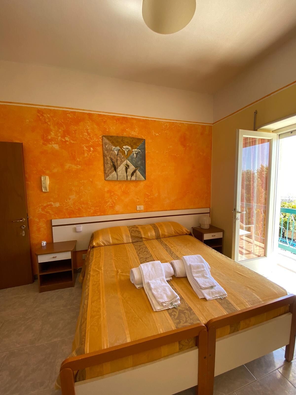 A bedroom with a bed, a painting on the wall, a nightstand, and a door leading to a balcony.