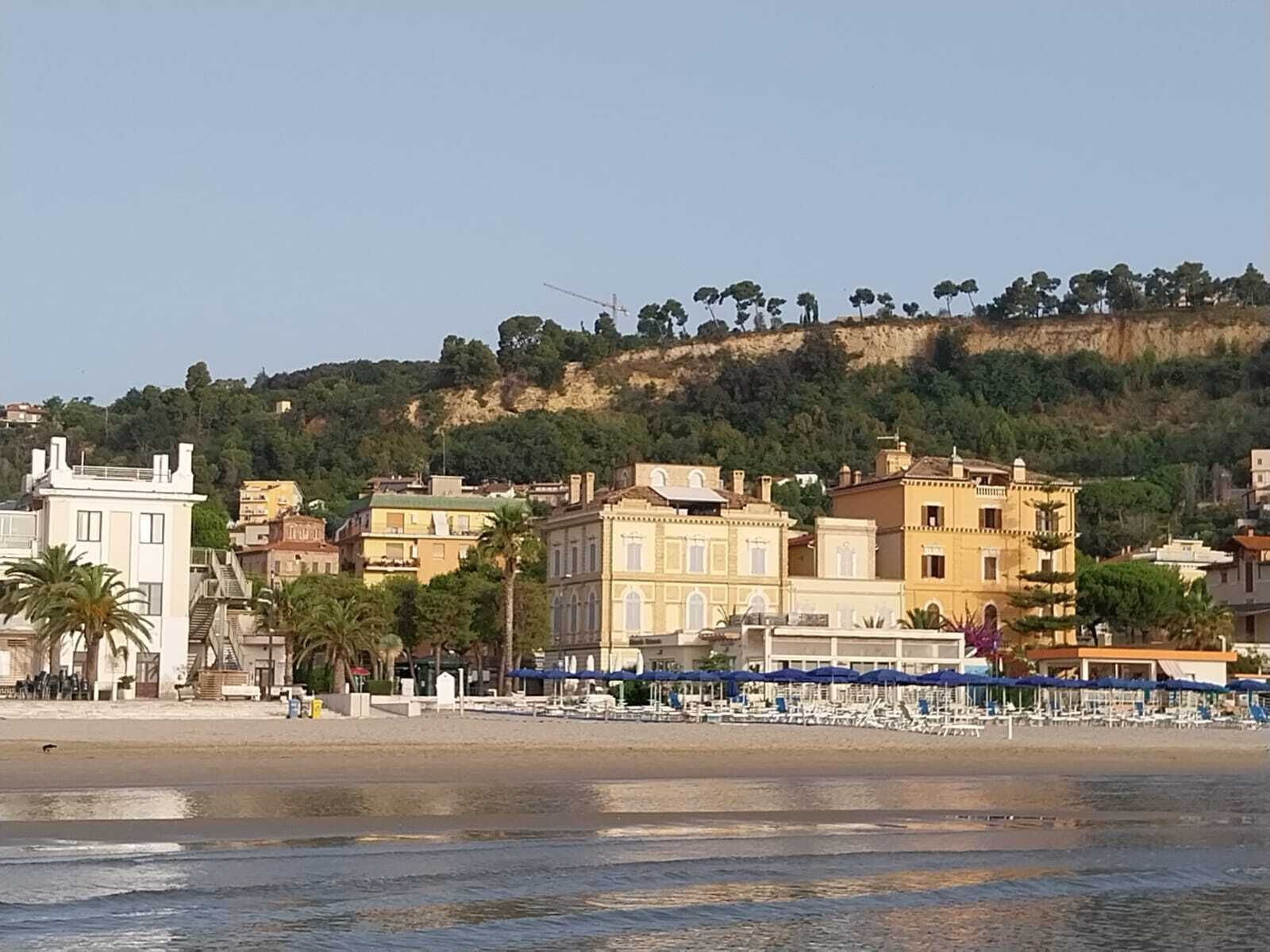 A coastal town with buildings along the beach, a clear sky, and a hilly backdrop.