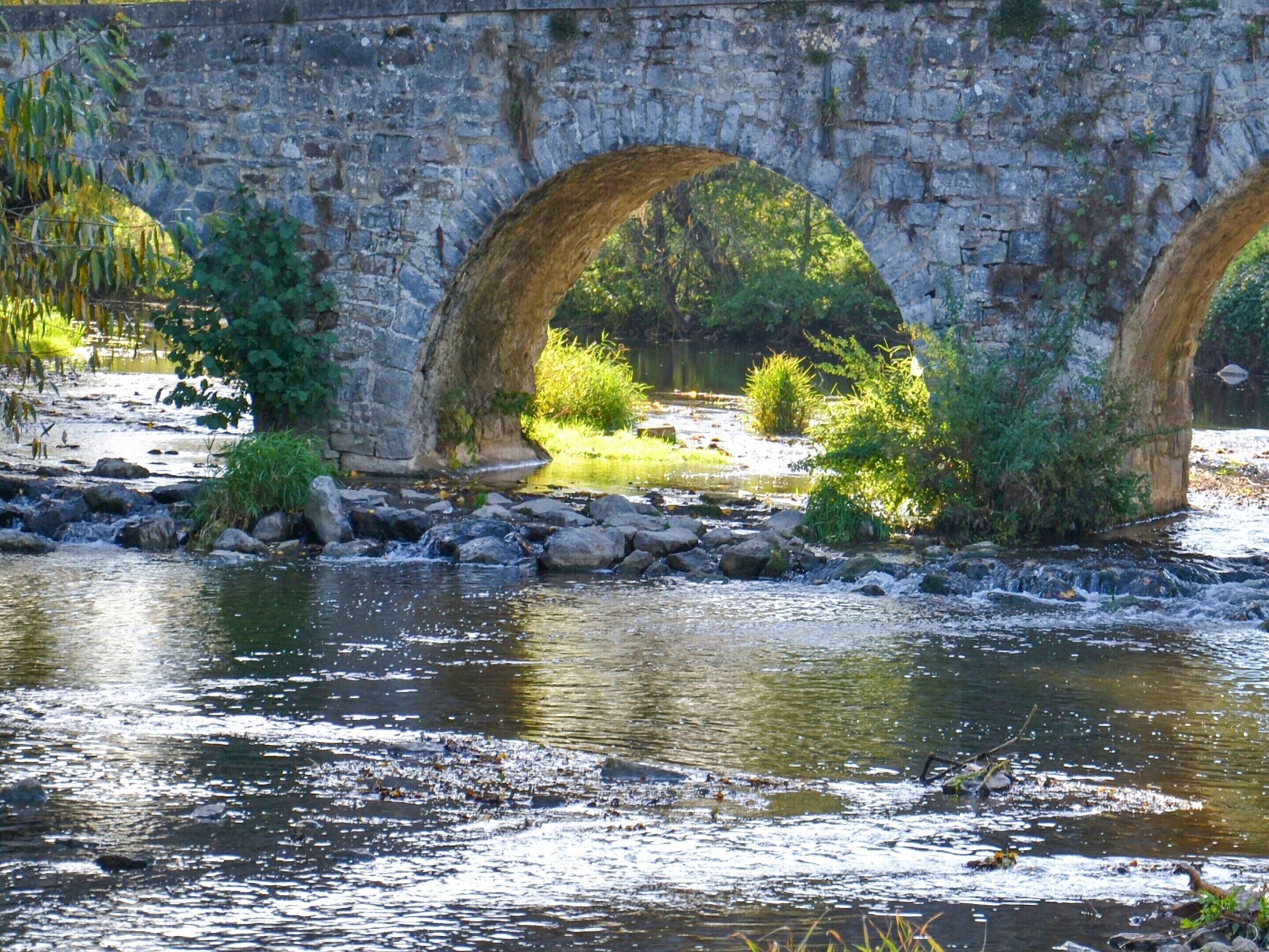 A stone bridge with three arches over a river, surrounded by greenery and rocks.