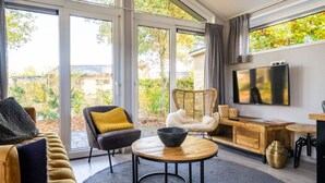 A modern living room with a sofa, a round coffee table, a wicker chair, and a flat-screen TV mounted on the wall. - Nice Chalet With Dishwasher, on the Veluwe (Otterlo)