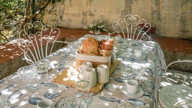 Ein Tisch im Freien mit Tischdecke mit Fischmuster, einem Brotkorb und mehreren Tassen und Gläsern.