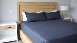 Une chambre à coucher avec une tête de lit en bois, un lit recouvert d’une literie bleu foncé, une table de chevet blanche avec un tiroir et une lampe posée sur la table de chevet.