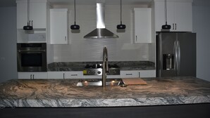 Une cuisine moderne avec des armoires blanches, une hotte de cuisinière en acier inoxydable, un four encastré, un réfrigérateur et des luminaires suspendus.