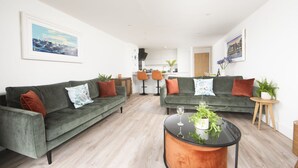 A modern living room with two green sofas, a round coffee table, and a kitchen in the background. - Sea Breeze -  a sea view apartment just moments from Lusty Glaze Beach (Newquay)