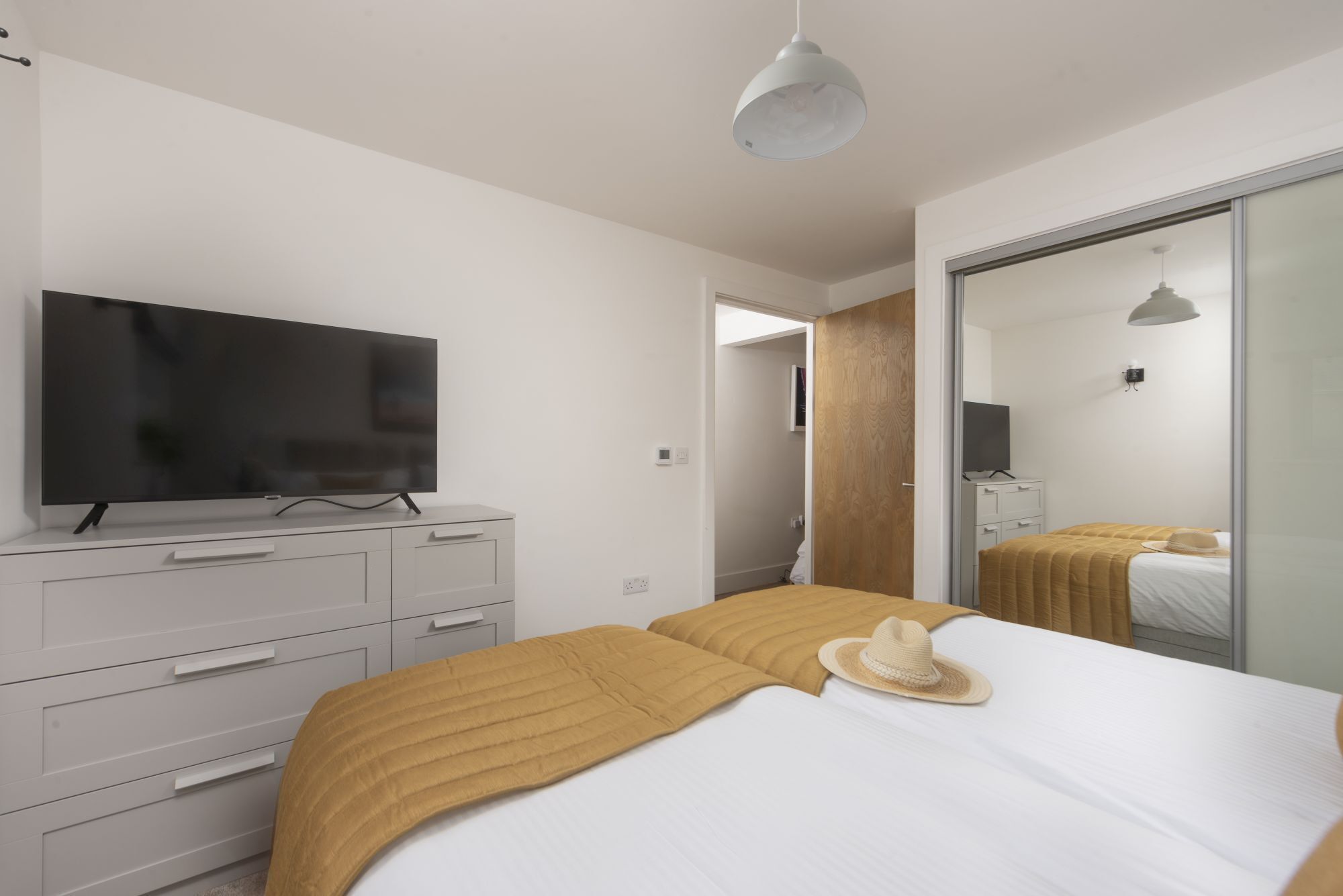 Une chambre moderne équipée d’un téléviseur à écran plat, d’une armoire, d’un lit avec une couverture beige et un chapeau, ainsi que d’un grand miroir.