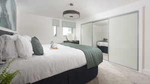A modern bedroom with a large bed, a bedside table, a mirror, and a sliding closet door. - Sea Breeze -  a sea view apartment just moments from Lusty Glaze Beach (Newquay)