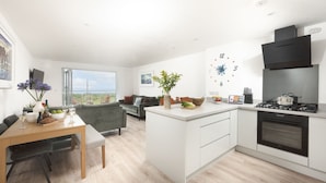 A modern kitchen with a central island, a dining area with a table and chairs, and a living space with a sofa and a view of the outdoors. - Sea Breeze -  a sea view apartment just moments from Lusty Glaze Beach (Newquay)