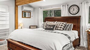 A bedroom with a wooden ceiling, a large bed with multiple pillows, a clock on the wall, and a window with curtains.