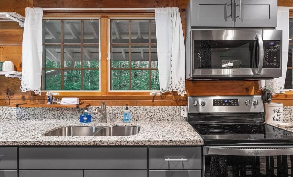 Una cocina con encimeras de granito, electrodomésticos de acero inoxidable, un lavamanos doble y una ventana con cortinas.