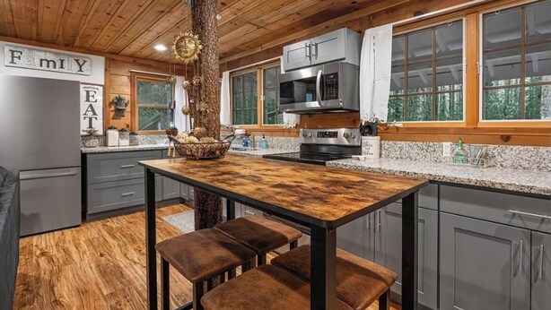 A modern kitchen with a wooden ceiling, grey cabinets, a central island, and a view of the outdoors through large windows.