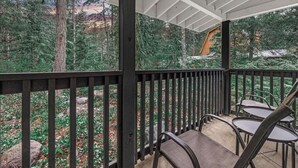 A covered outdoor area with a wooden railing, seating, and a view of a forested landscape.