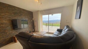 A living room with a large L-shaped black leather sofa, a flat-screen TV mounted on a brick wall, and a sliding glass door leading to a patio with a view of the countryside.