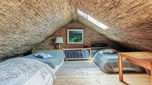 An attic bedroom with a thatched ceiling, a bed with patterned bedding, a small table, a lamp, and a painting on the wall.
