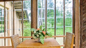 A wooden-framed room with a table and two chairs, a vase with flowers, and a view of greenery through large windows.
