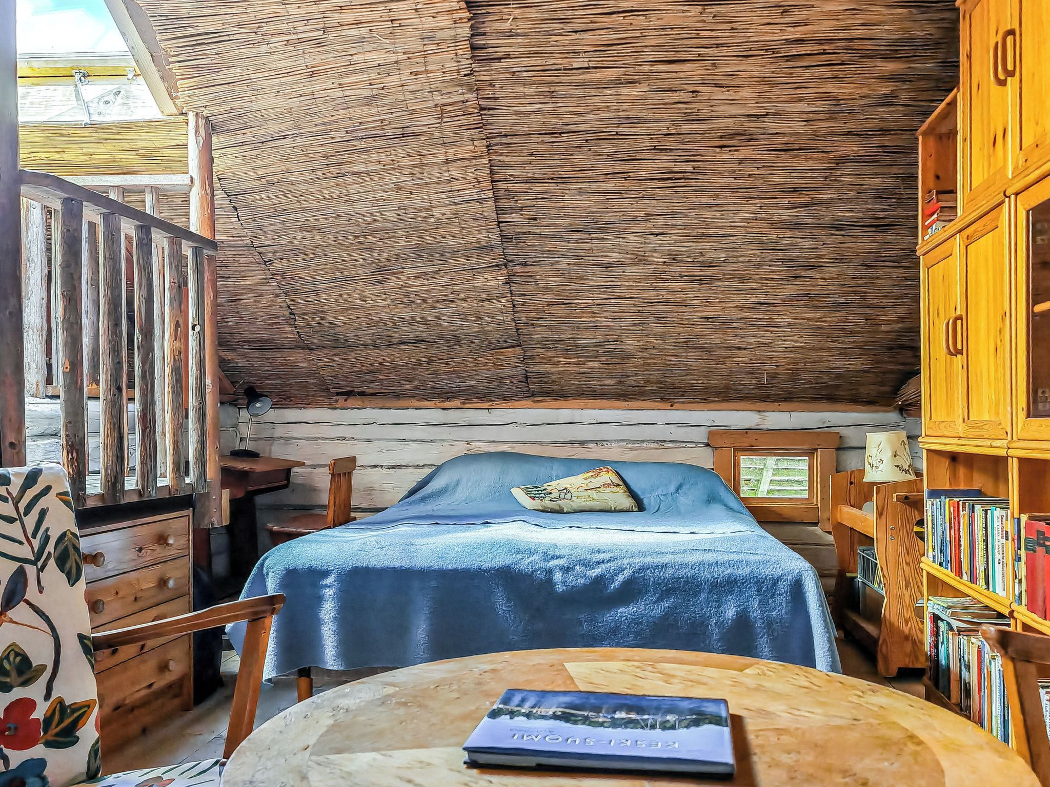 A small, cozy room with a bed, a wooden cabinet with books, and a table with a book on it.