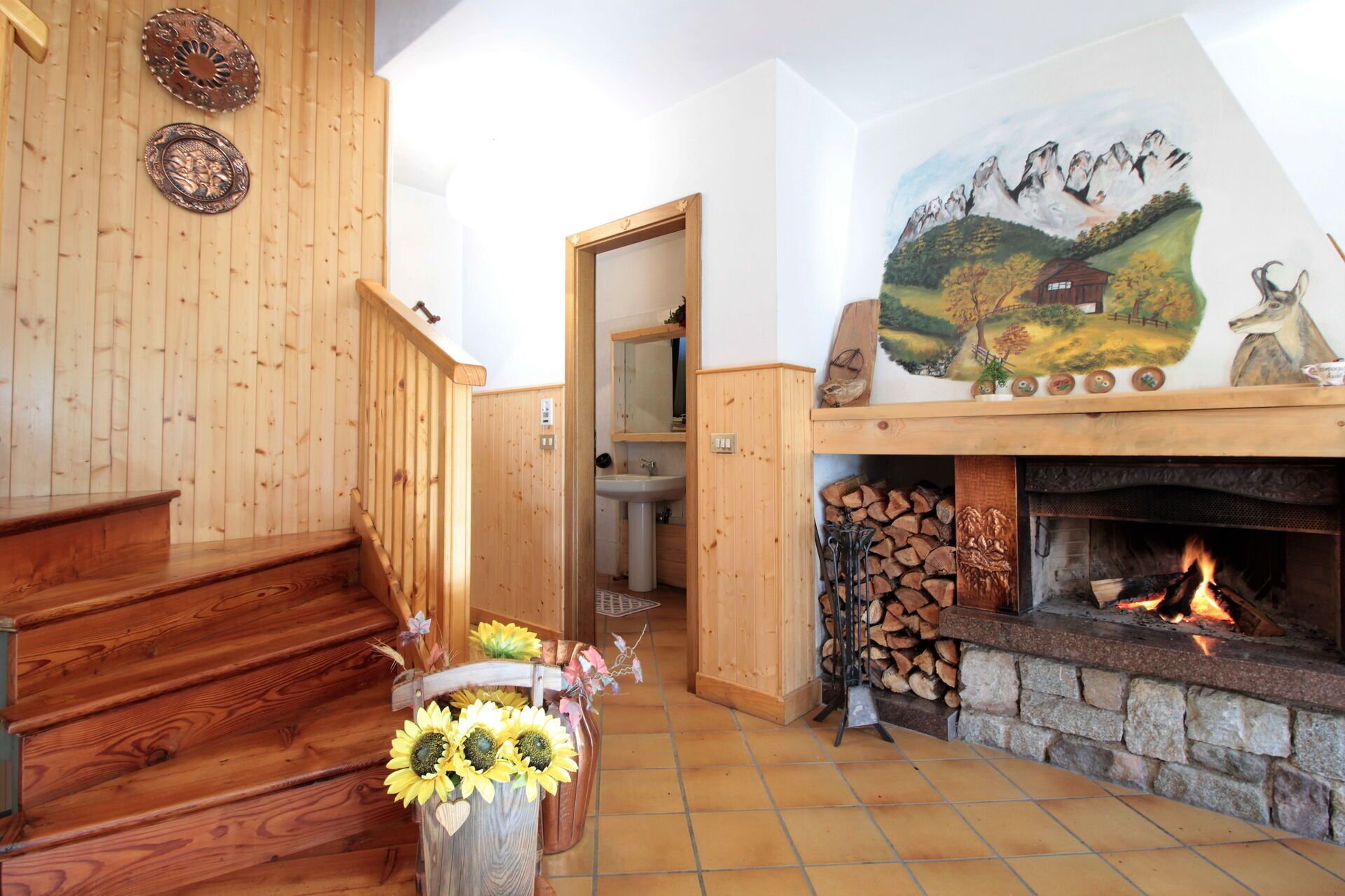 A wooden staircase leading to a bathroom with a sink and mirror. A fireplace with a stone surround and a painting of a mountain scene is visible.