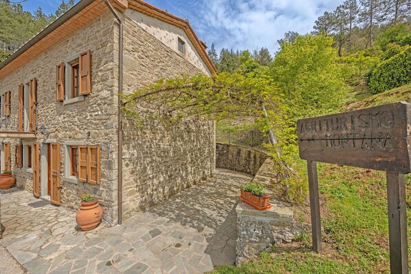 A stone building with wooden shutters, a sign indicating 'Agriturismo Montagna', and potted plants.
