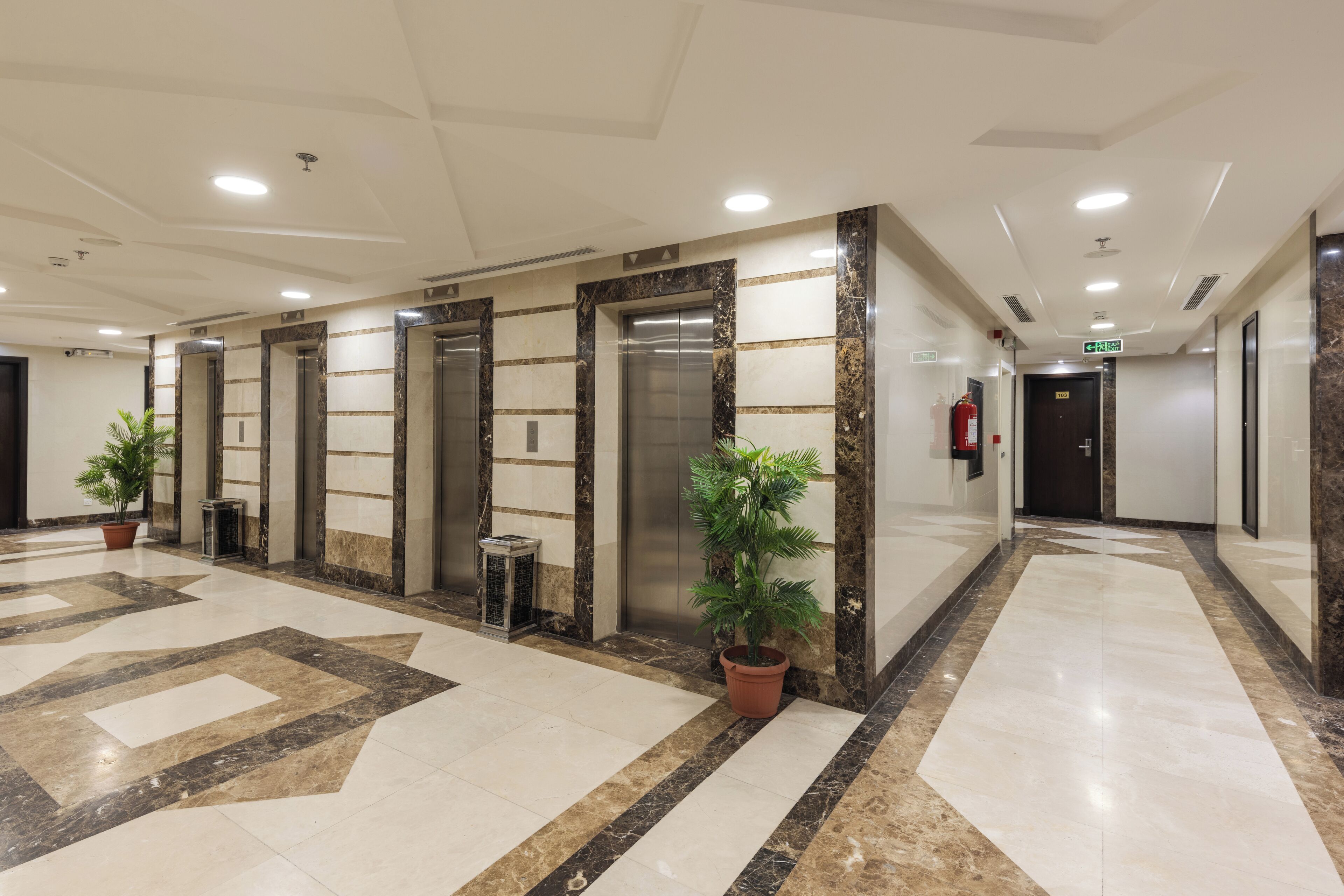 A modern hotel lobby with marble flooring, elevators, potted plants, and a fire extinguisher.