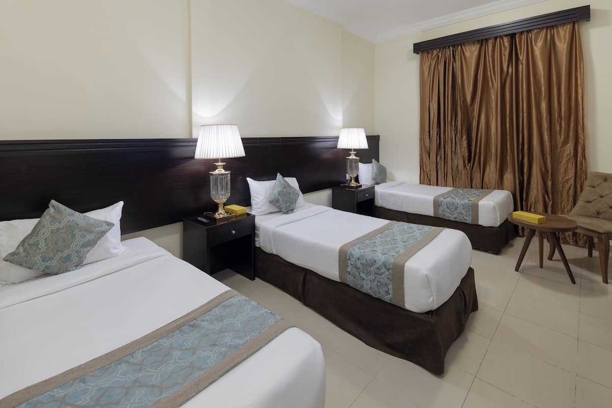 Une chambre d’hôtel avec trois lits simples, chacun doté d’une table de chevet, d’une lampe et de coussins décoratifs. La chambre dispose d’une grande fenêtre avec des rideaux, d’une tête de lit en bois et d’une petite table ronde avec une chaise.