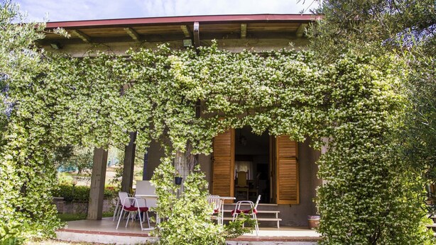 Un bâtiment doté d’un porche abrité, de volets en bois et d’un petit jardin agrémenté de chaises et de tables.