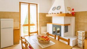 Une cuisine équipée d’un réfrigérateur, d’une table en bois et de chaises. On y trouve une fenêtre avec des rideaux, un radiateur et une petite étagère ornée d’objets décoratifs.