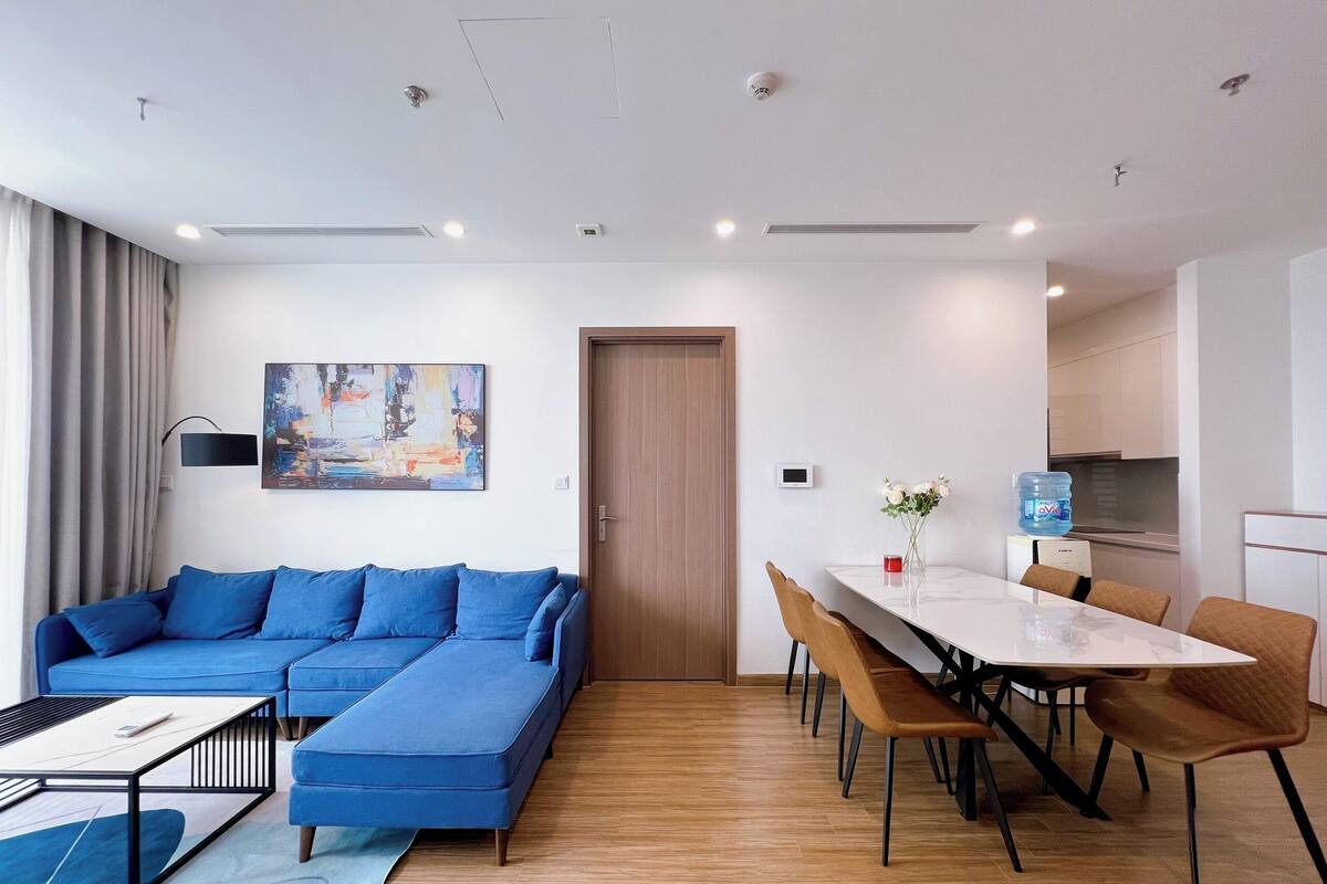 A modern living room with a blue sectional sofa, a dining table with chairs, and a kitchen area in the background.