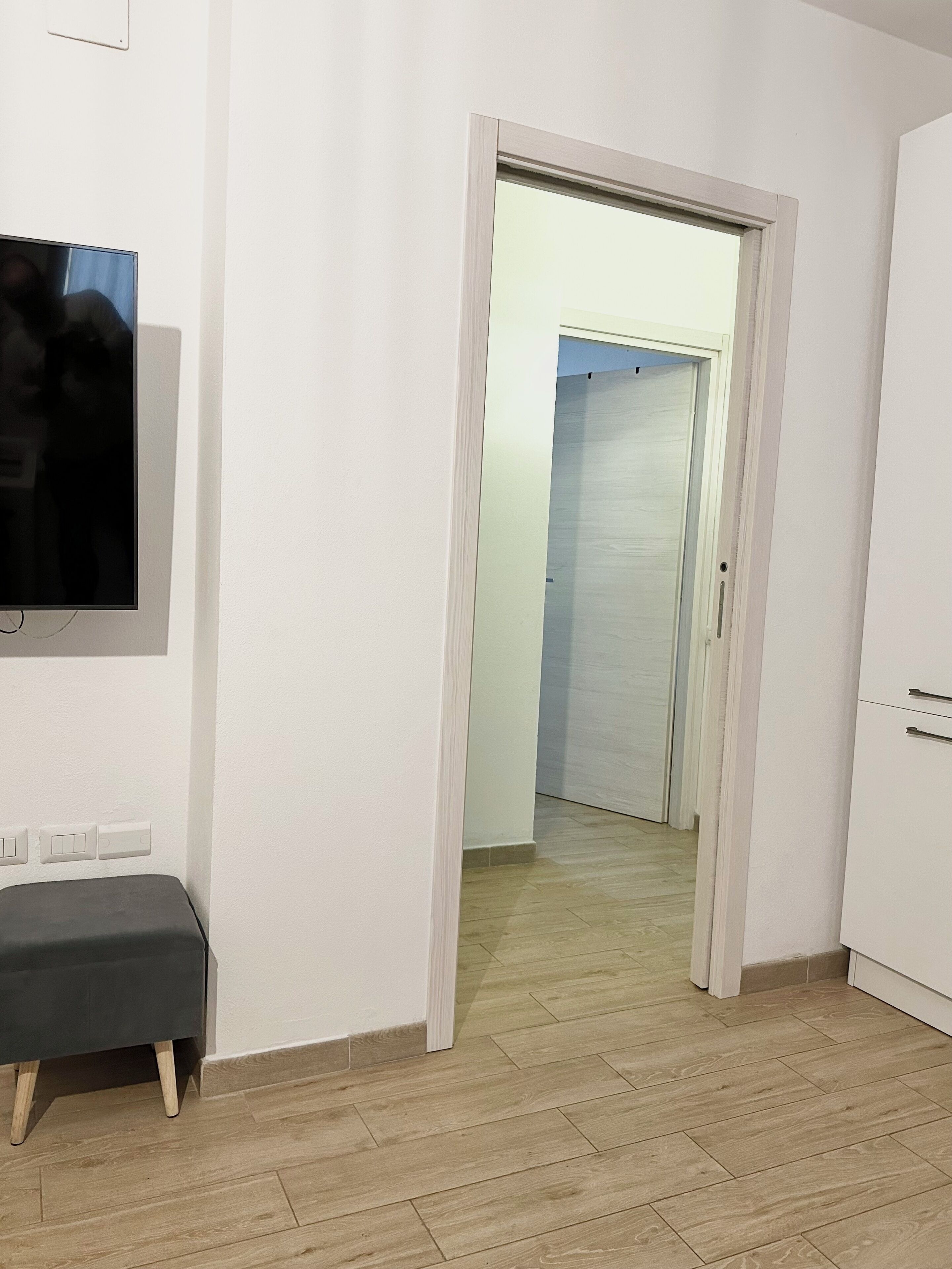 A room with a flat-screen TV mounted on the wall, a bench, and a door leading to another room.