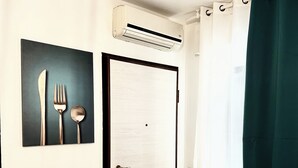 A room with a white door, an air conditioning unit above it, a window with a green curtain, and a wall-mounted picture of cutlery.