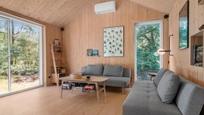 A cozy cabin interior with wooden walls, a grey sofa, a wooden table with toys, and a large window offering a view of trees. - 3 bedroom amazing home in Stege (Stege)