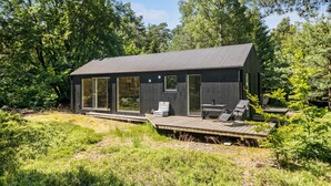 A modern cabin with large sliding glass doors, a deck with outdoor seating, and a forested backdrop. - 3 bedroom amazing home in Stege (Stege)
