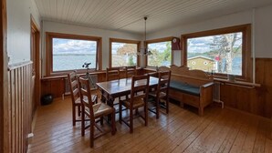 A wooden cabin with a dining table and benches, large windows overlooking a lake, and a view of a building across the water. - 4 bedroom amazing home in Sorunda (Sorunda)