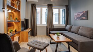 A modern living room with a sofa, a small table, a TV, and a bookshelf with decorative items. - Agora Apartments (Lesvos)