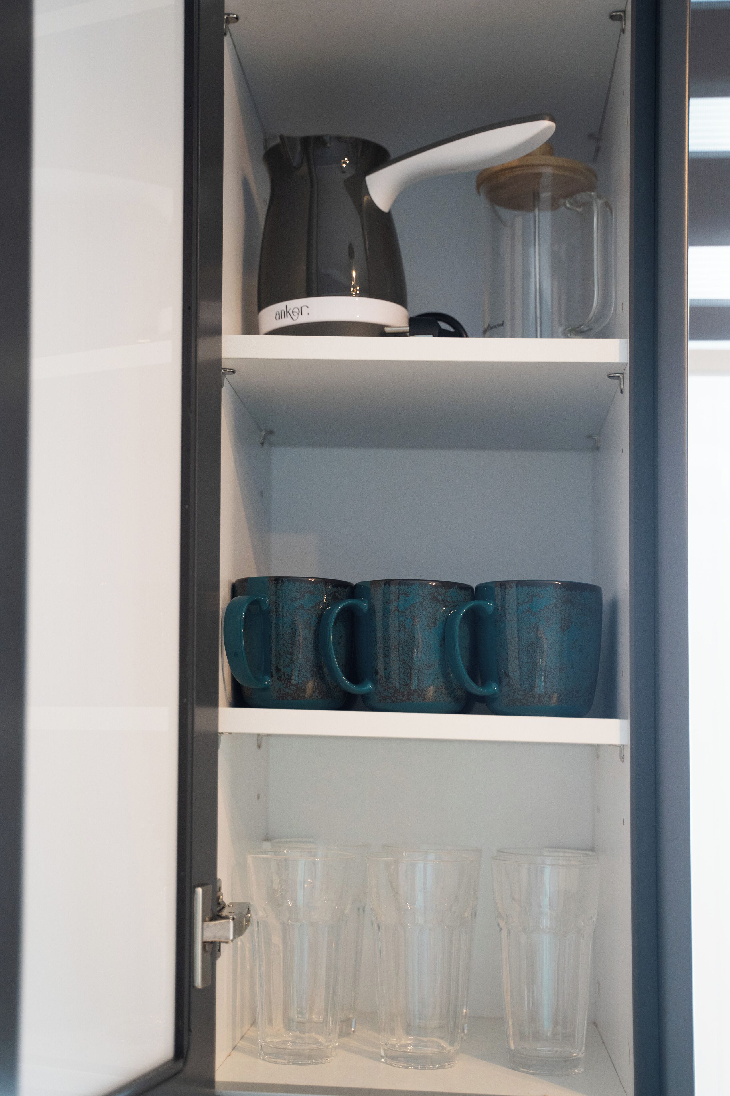 Una cocina con cafetera, un tarro y varias tazas y vasos en estantes.