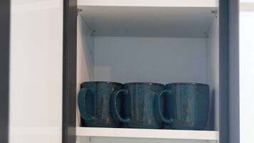 Una cocina con cafetera, un tarro y varias tazas y vasos en estantes.