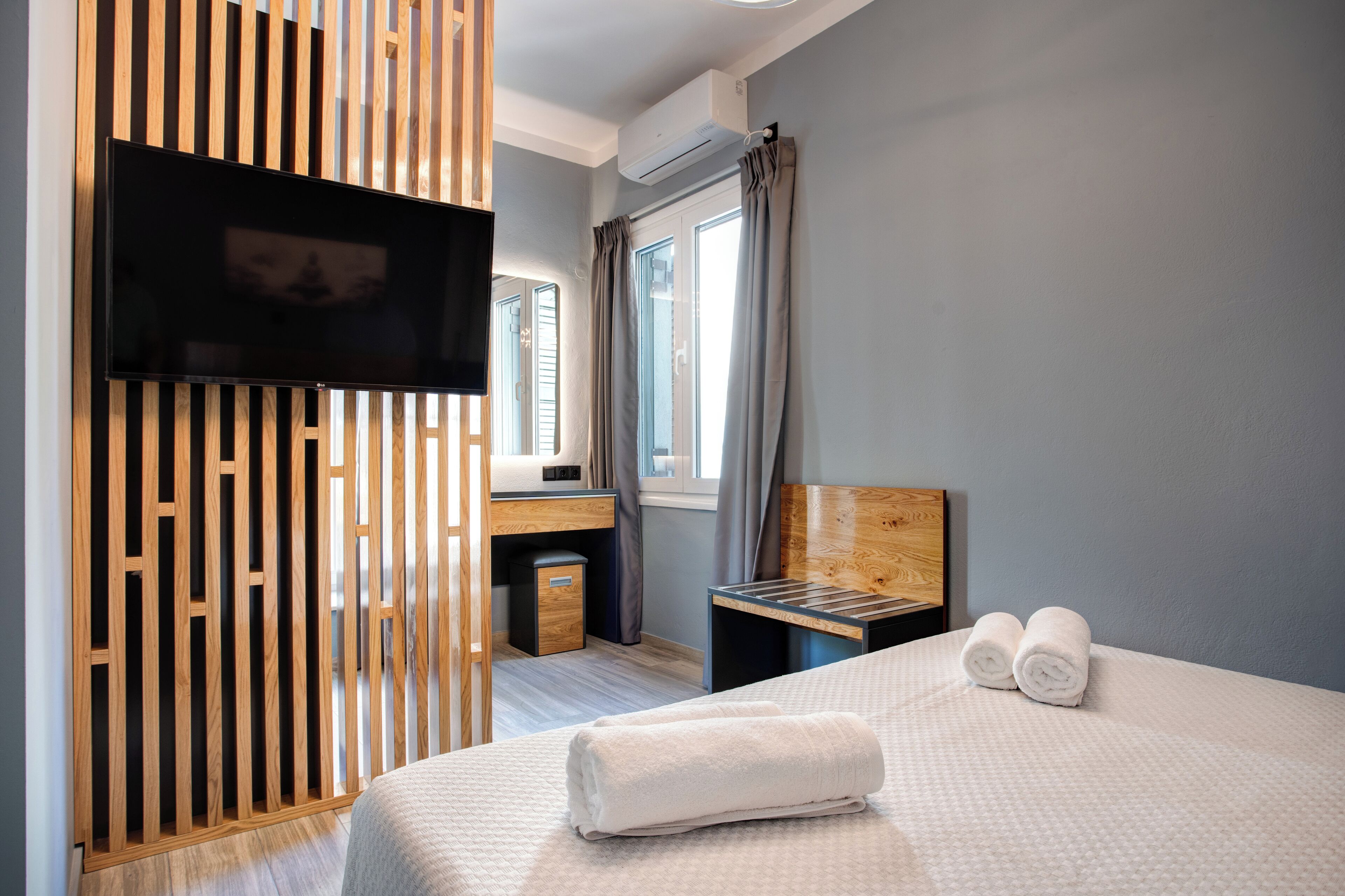 Una habitación de hotel con una cama, un televisor montado en un panel de madera, un escritorio con una silla y una ventana con cortinas.