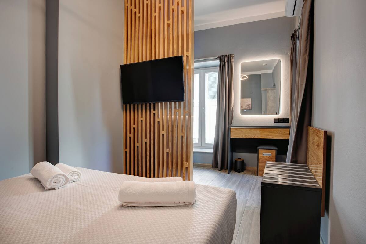 Une chambre d’hôtel moderne avec un lit bien fait, un téléviseur fixé sur un panneau en bois, une salle de bain avec un miroir et une petite table de chevet.