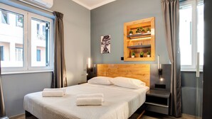 A modern hotel room with a large bed, a wooden headboard, a wall-mounted shelf with plants, and a window with curtains. - Agora Apartments (Lesvos)