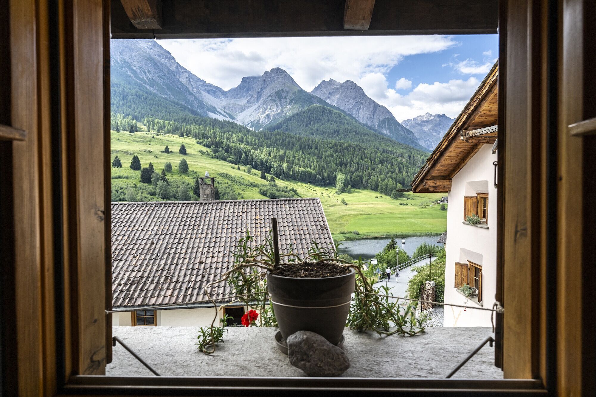 A window view of a mountainous landscape with a house and a potted plant on the windowsill.