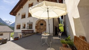 Ein Innenhof mit Tisch und Stühlen unter einem großen Sonnenschirm, einer hölzernen Bank und Blumentöpfen.