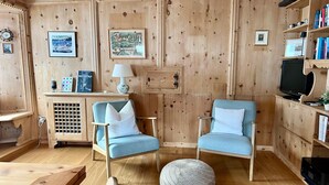 Ein gemütlicher Raum mit Holzwandverkleidung, zwei blaue Sessel mit weißen Kissen, ein runder gewebter Hocker, ein Holzschrank mit Lampe sowie ein Fernseher auf einem Holzgestell.