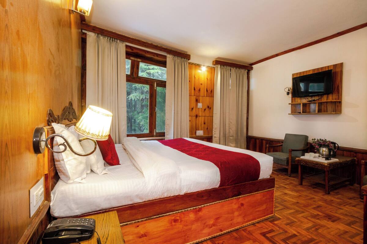 Una cama con estructura de madera y ropa de cama blanca y roja, una lámpara de noche, un teléfono y un televisor montado en la pared.