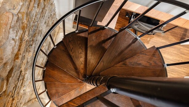 A spiral staircase with wooden steps and metal railings, leading to a room with a wooden floor and a stone wall. - 4BR House | City Views | Balconies | Walk to OTR (Cincinnati)