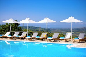 Zona de piscina con sillones de descanso y sombrillas, cielo azul despejado y vistas al paisaje.