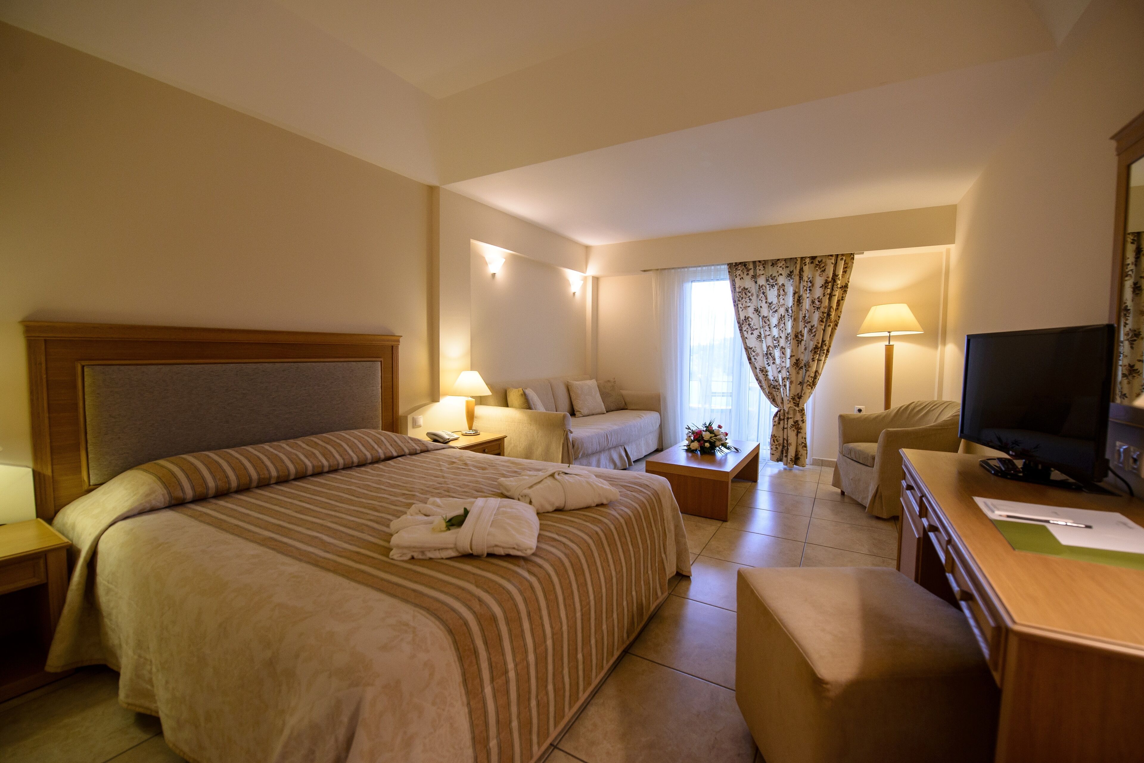 Une chambre d’hôtel avec un grand lit, un canapé, un bureau avec une télévision et une fenêtre avec des rideaux.