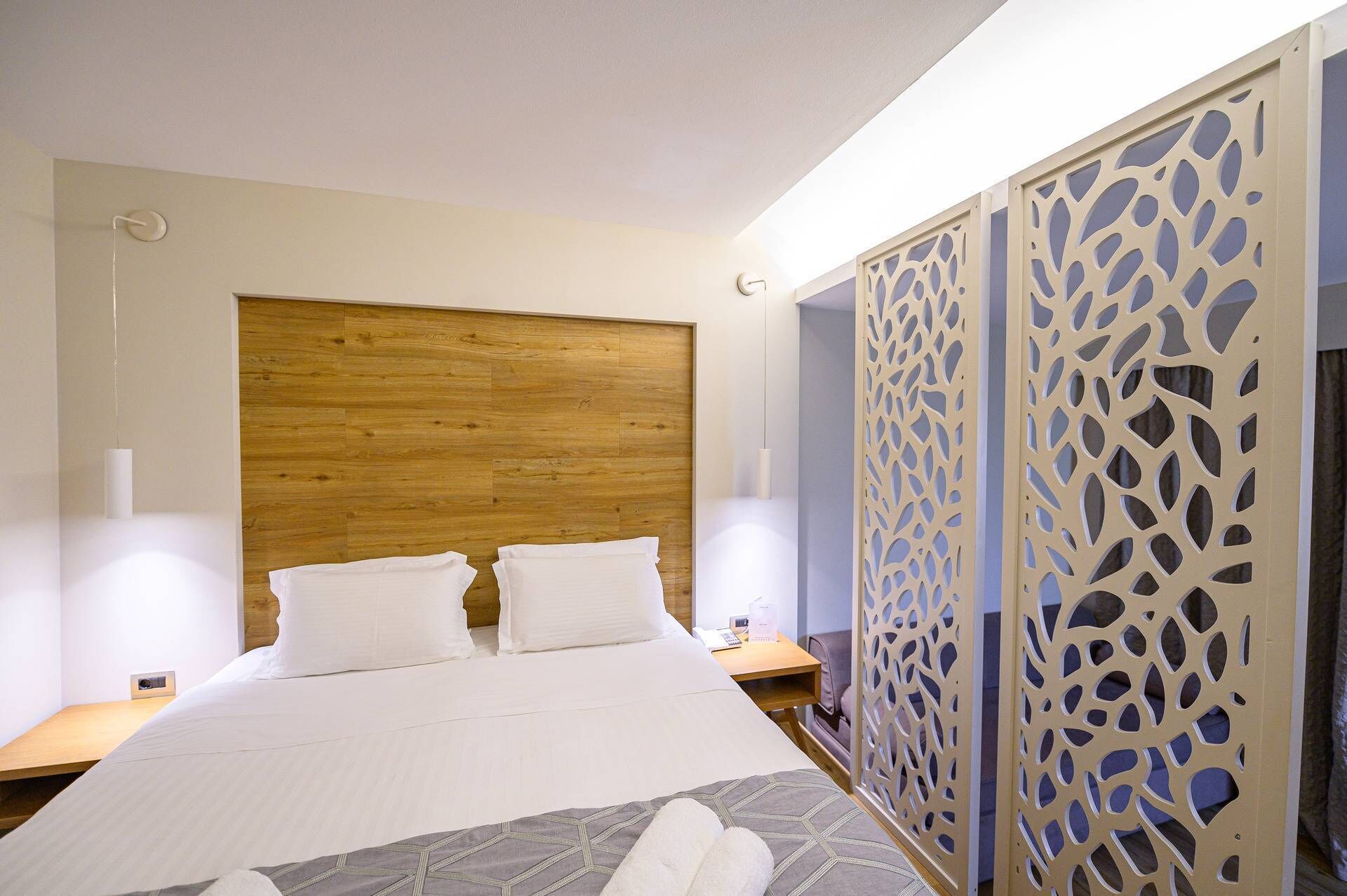 Une chambre d’hôtel avec une tête de lit en bois, un lit recouvert de draps blancs, deux tables de chevet et un paravent décoratif orné de motifs découpés.
