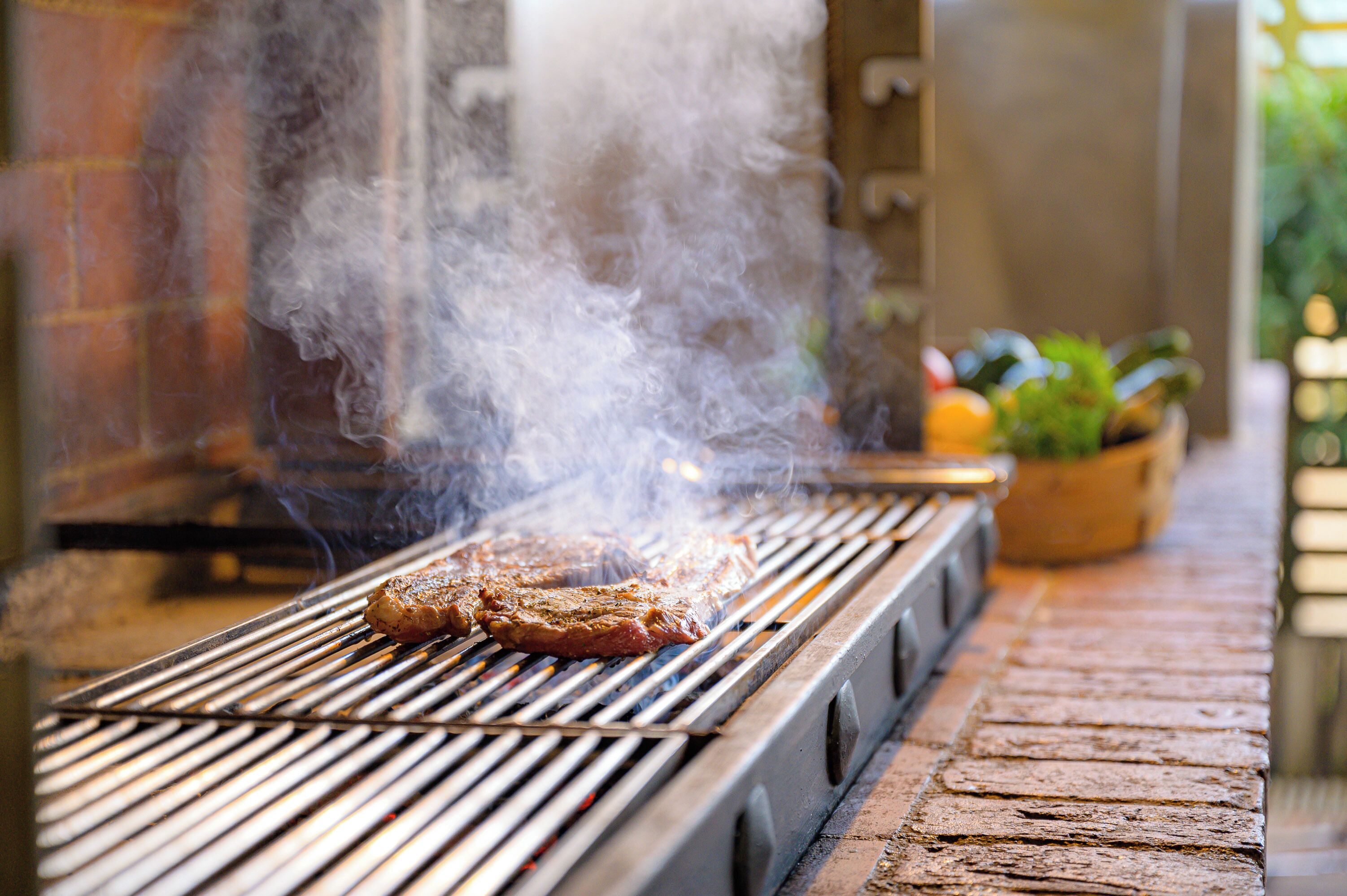 A grill with two steaks cooking, emitting smoke, and a brick wall in the background.