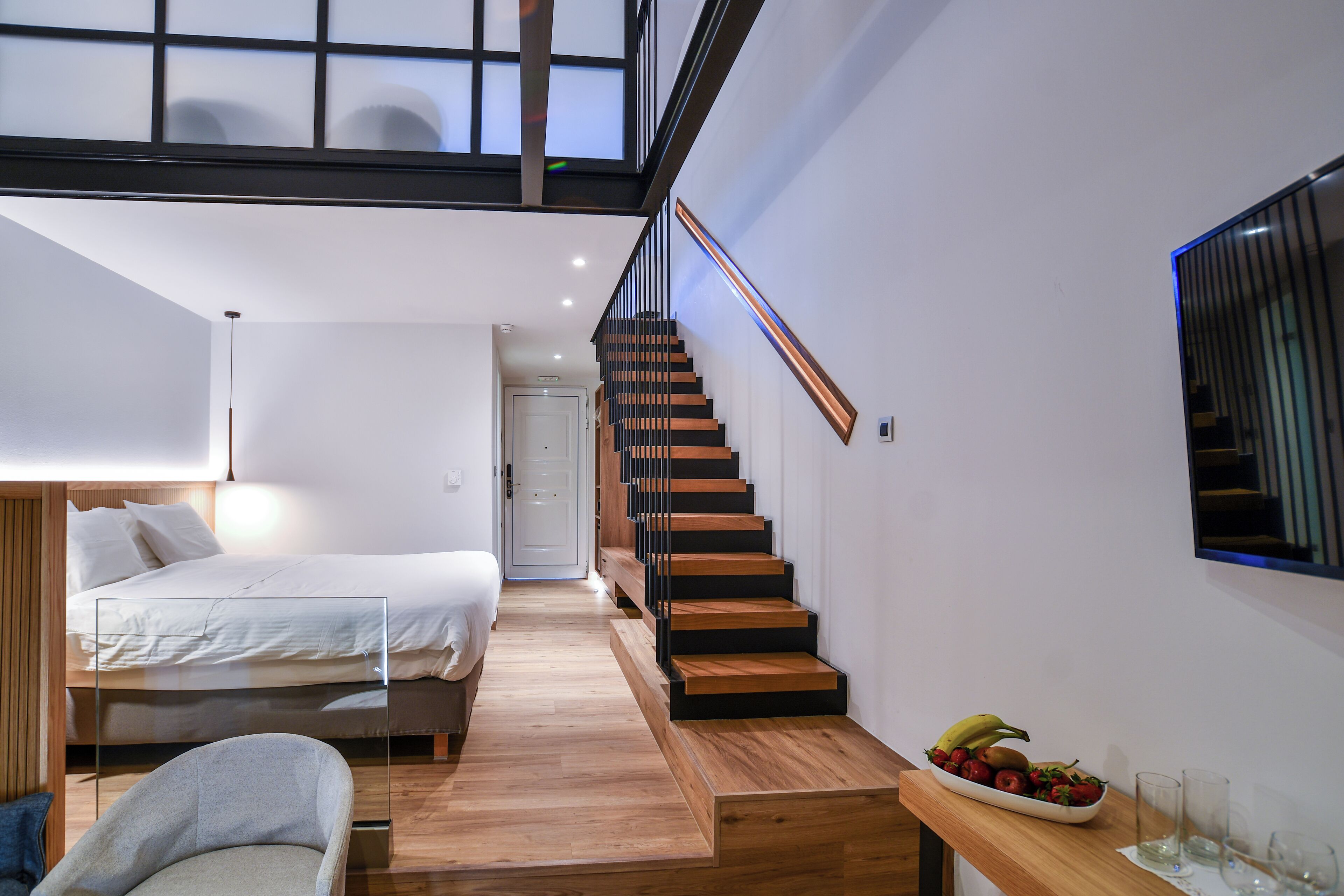 Un dormitorio moderno con una escalera de madera, una cama, una mesita de noche con un frutero y un televisor montado en la pared.