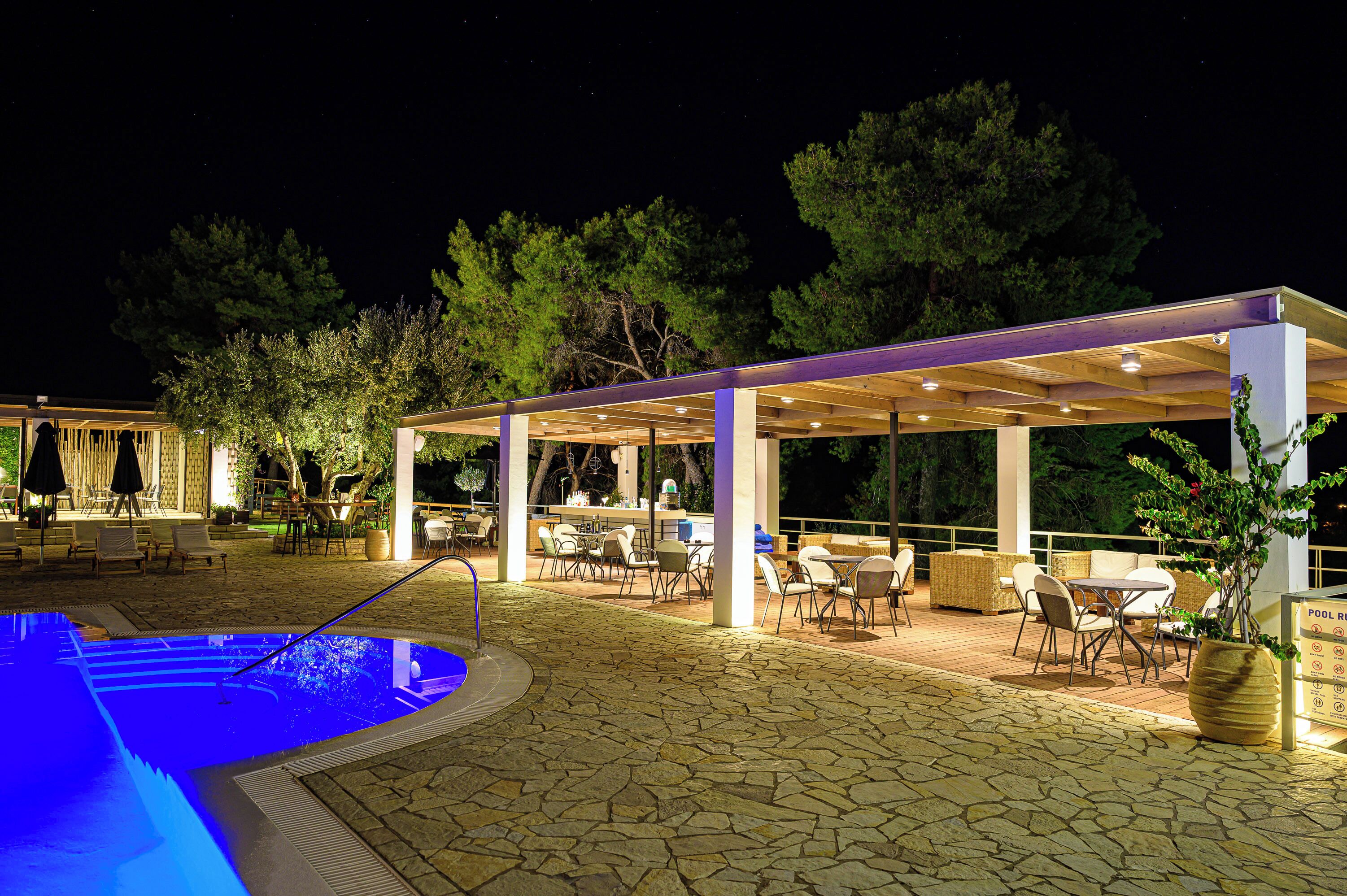 An outdoor pool area with a covered seating area, stone flooring, and surrounding trees.